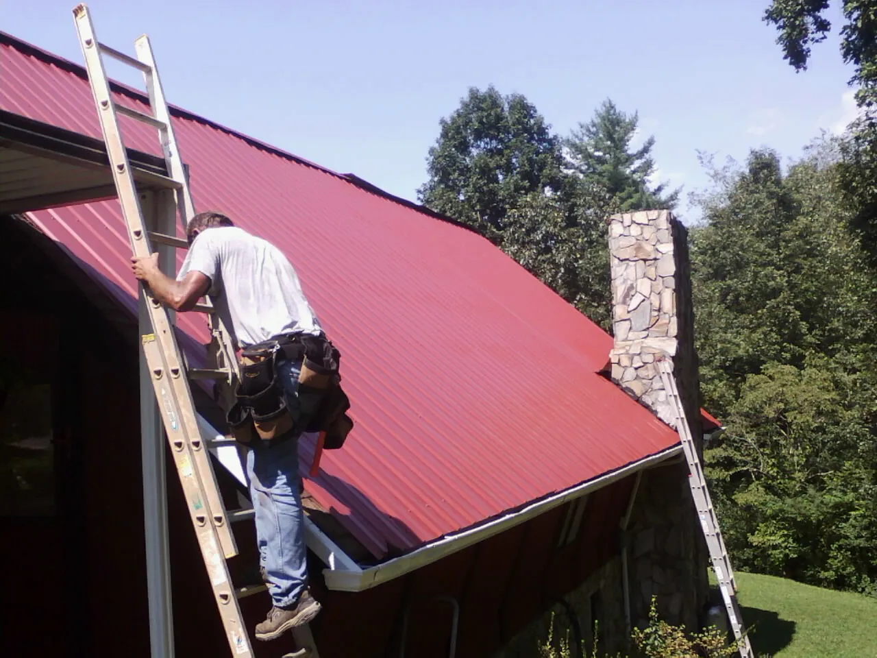 Roofer on a ladder beside a red metal roof and stone chimney — Soffit & Fascia Repair by Roofing Brothers in Blacksburg